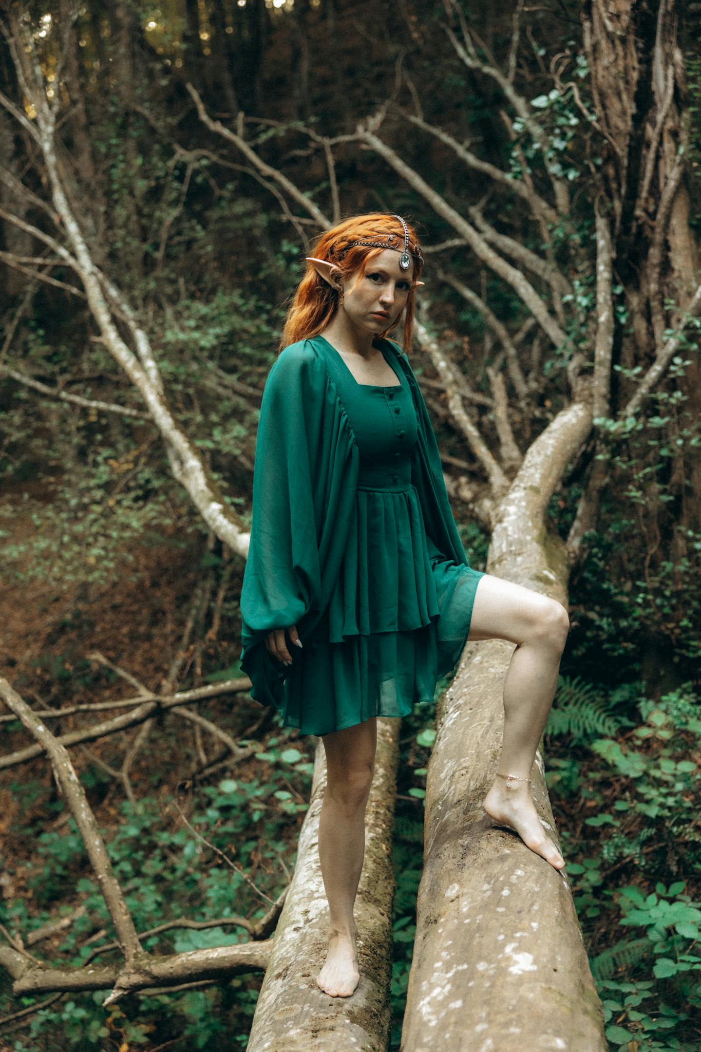 A woman in green fantasy attire poses on a log in a mystical forest.