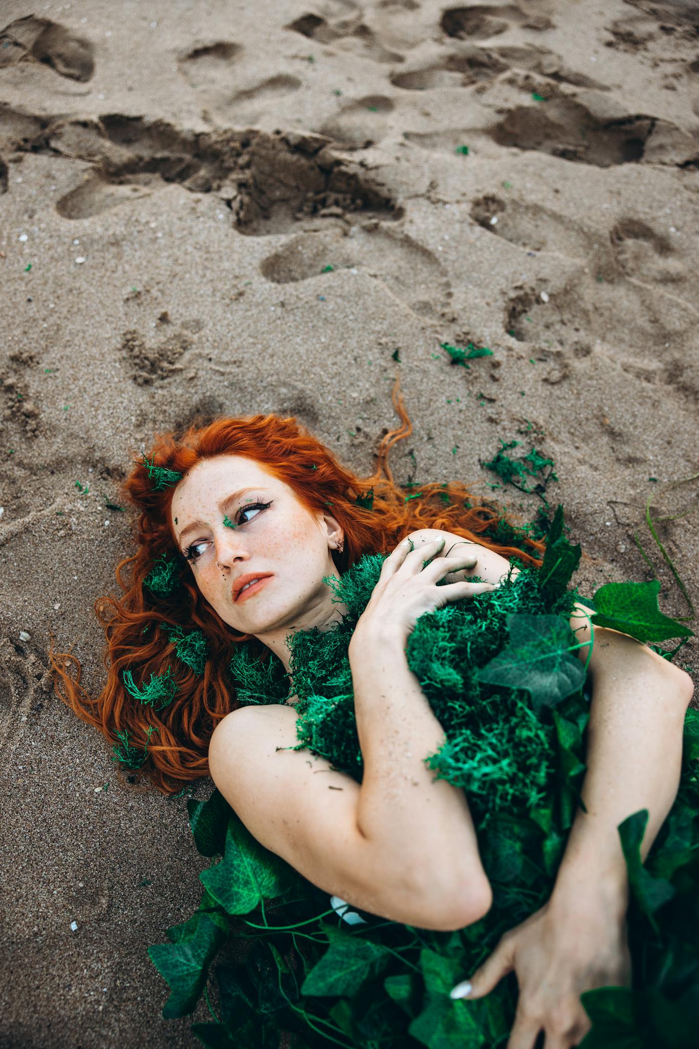 Red-haired woman with greenery artfully draped, lying on sandy beach.
